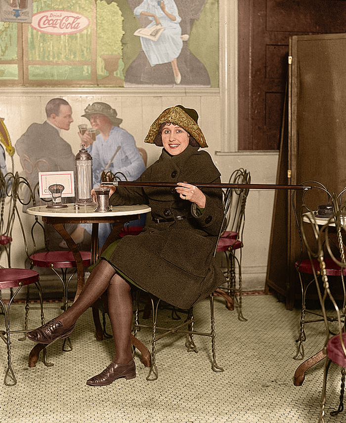 Woman seated at a soda fountain table pours alcohol into her cup from a cane. Note the large Coca-Cola advertisement on the wall,