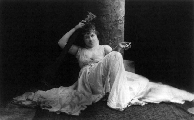 Loie Fuller, full length, seated, facing front; holding cigarette and stringed musical instrument.Source
