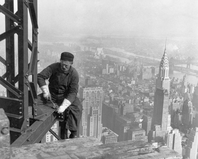 A worker bolts beams during construction; the Chrysler Building can be seen in the background. Source