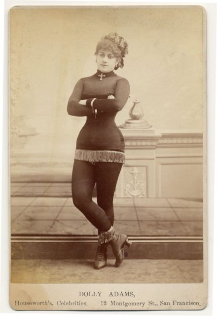 Dolly Adams with fringe at the bottom of a short costume, tights, short-heeled boots topped with fringe, cross at neck, cap.