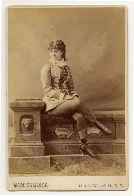 Minnie Marshall sitting on a stone fence, in a short dress and boots.