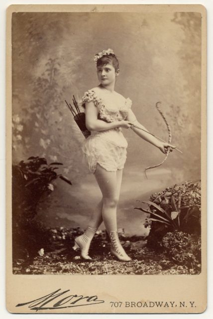 Miss Farrington in a short sleeveless costume, holding bow and arrow, with quiver and arrows on back, buttoned and heeled boots.