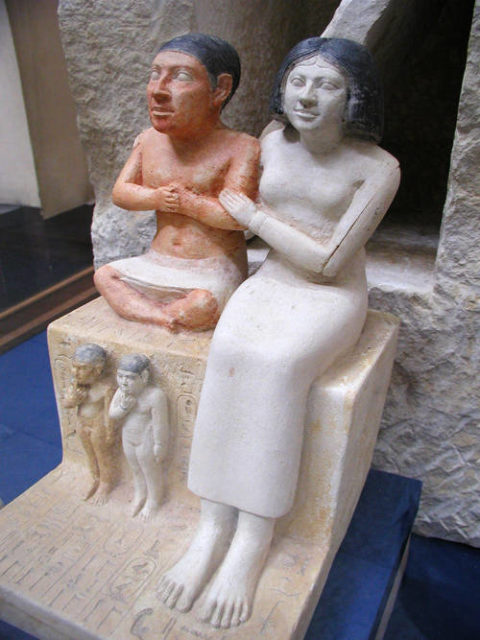 The limestone sculpture of Seneb and his family is part of the collection of the Egyptian Museum in Cairo. It depicts Seneb and his wife sitting next to each other with their children in the lower register. Seneb is depicted sitting cross-legged on a block of stone with his arms folded in a position characteristic of a scribe. His wife Senet sits alongside him, wearing a long robe with long sleeves and a wig covering her natural hair, which can be glimpsed on her forehead. She encircles him with her arms in a gesture of affection and support. She is shown with a slight smile on her face to signify her contentment and happiness. By Jon Bodsworth - http://www.egyptarchive.co.uk/html/cairo_museum_12.html, Copyrighted free use, https://commons.wikimedia.org/w/index.php?curid=3786467