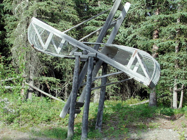 A wooden fish wheel out of the water. This wheel would rotate clockwise as water flowed from right to left. Openings on the near side, close to the axle, are where fish would pour out, propelled by the slanted bottom of the basket when it is nearing the top of the wheel. Wikipedia Public Domain
