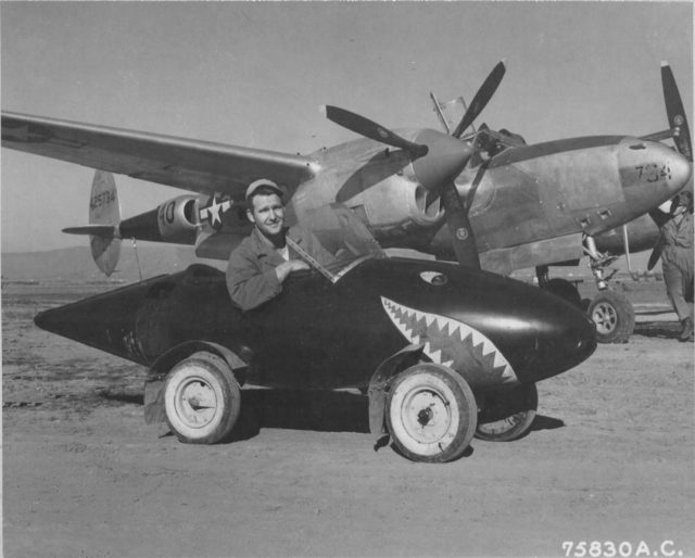 United States Army Air Forces Lockheed P-38L Lightning aircraft ( Serial Number - 44-25734 ) and a ground crew member of the 94th Fighter Squadron 1st Fighter Group, poses in his self-styled auto made from salvaged Lockheed P-38 Lightning parts including a fuel tank with wheels added and a plexiglass windshield. This P-38 while assigned to 1st FG, 71st FS was shot down by AAA near Munich April 15, 1945
