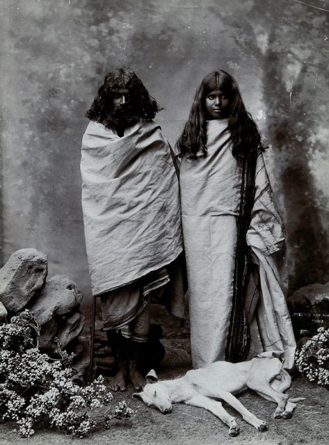 A man and woman of the Toda tribe standing in a photographic studio, with a dog lying at their feet. Photograph, ca.1900. Iconographic Collections