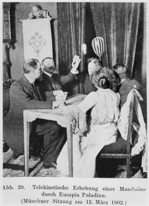 Mandolin (striped instrument, top, right) levitates above Palladino's head in front of the curtains at the far short end of the table during Palladino's séance in Munich, Germany, 13 March 1903