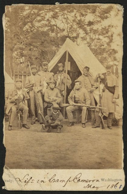 Photograph shows uniformed soldiers with guns posed in front of a tent; an African American child sits in front of the group. Photo Credit