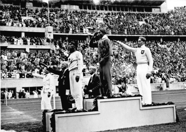 Jesse Owens on the podium after winning the long jump at the 1936 Summer Olympics. L–R, Naoto Tajima, Owens, Lutz Long. Photo by Bundesarchiv, Bild 183-G00630 / Unknown / CC-BY-SA 3.0.