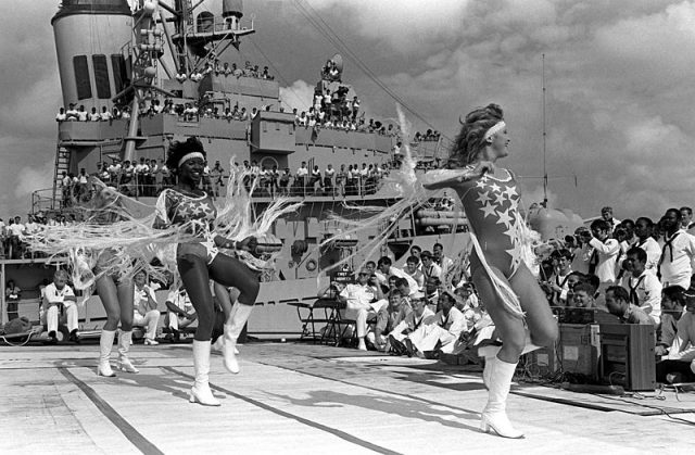 : The Dallas Cowboys Cheerleaders perform their USO show “America and Her Music” on the deck of the nuclear-powered guided missile cruiser USS Bainbridge (CGN-25). Crew members of the guided missile destroyer USS Waddell (DDG-24) watch from their ship. 1983