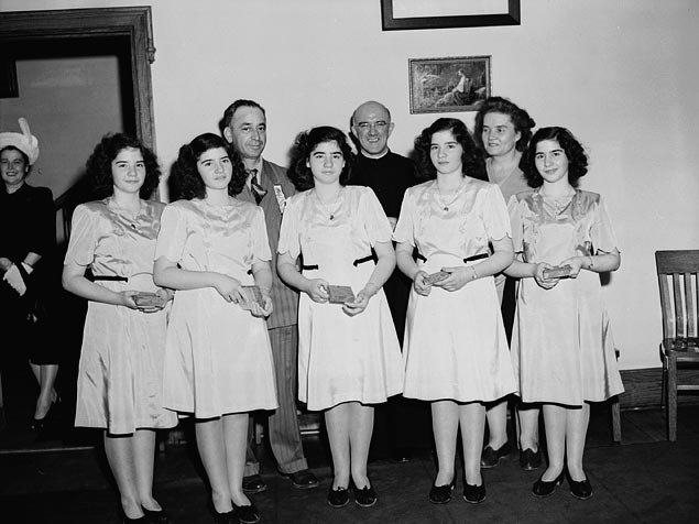 The quintuplets in 1947 with their parents and a priest in the background.