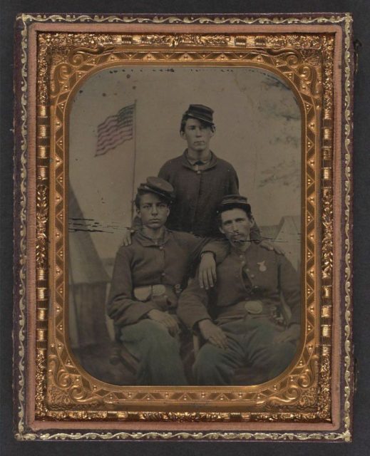 Three unidentified Union soldiers in front of painted military camp scene backdrop.