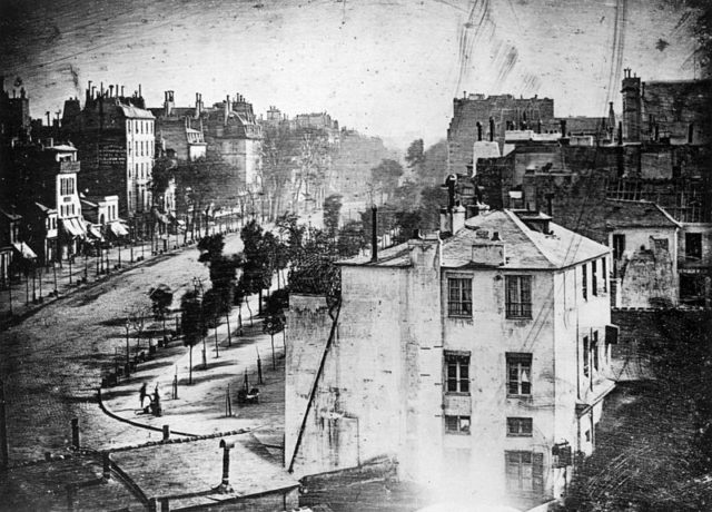 The earliest reliably dated photograph of people, taken by Daguerre one spring morning in 1838 from the window of the Diorama, where he lived and worked. It bears the caption huit heure du matin (8 a.m.). Though it shows the busy Boulevard du Temple, the long exposure time (about ten or twelve minutes) meant that moving traffic cannot be seen; however, the bootblack and his customer at lower left remained still long enough to be distinctly visible. The building signage at the upper left shows that the image is laterally (left-right) reversed, as were most daguerreotypes. Daguerre presented this daguerreotype together with two others: a still-life and a view from the same window labelled midi (noon) to King Ludwig I of Bavaria (The Munich Triptych) in order to publicise his invention. All three daguerreotypes were destroyed by cleaning in 1974 but they are preserved in reproduction. Wikipedia/Public Domain