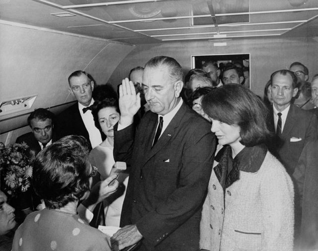 Kennedy, still wearing the blood stained pink Chanel suit, stands alongside as Lyndon B. Johnson takes the Presidential oath of office aboard Air Force One. Wikipedia/Public Domain