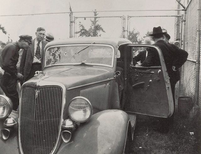 Highway Patrolman on left beside Sheriff Smoot Schmid,inspects the Barrow “death car” while Frank Hamer (on the right, with his back to the camera) talks with other officials. 