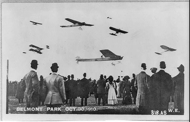 Crowd watching seven planes in air at Belmont Park air show, New York, year 1910. 