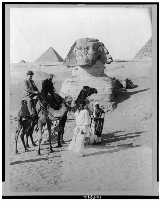 Two men on camels and two Egyptians in front of the Sphinx, with pyramids in the background, Giza, Egypt. Photo Credit 