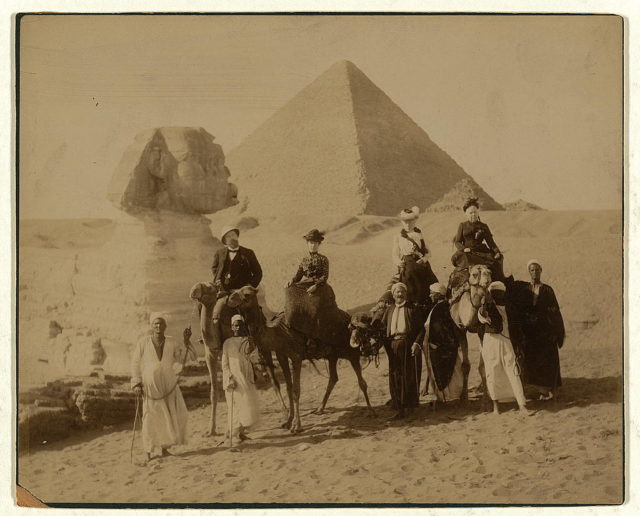 Three women and a man in western attire, seated on camels, several local men stand before the camels holding the reins, a pyramid and the sphinx in the background Photo Credit 