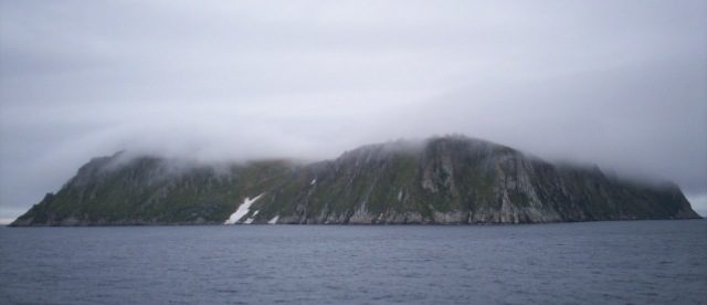 King Island, Alaska. The large boulders on the top of the island are barely visible through the fog. The abandoned village is in the vicinity of the snow drift. Photo credit