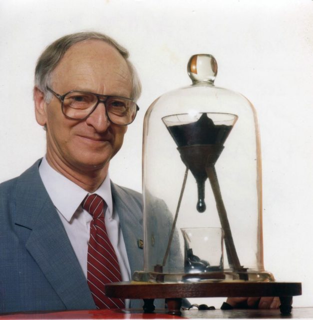 The University of Queensland pitch drop experiment, featuring its previous custodian, Professor John Mainstone (taken in 1990, two years after the seventh drop and 10 years before the eighth drop fell). – John Mainstone, University of Queensland – CC BY-SA 3.0
