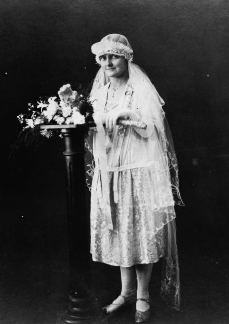 The bride is wearing a blouson top wedding dress with calf-length full lace skirt, her veil’s headpiece is surrounded by a tulle ruffle