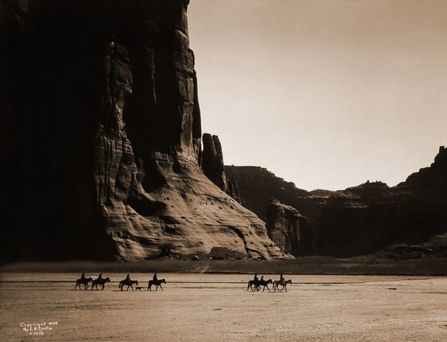 Canyon de Chelly – Navajo. Seven riders on horseback and dog trek against background of canyon cliffs, 1904.