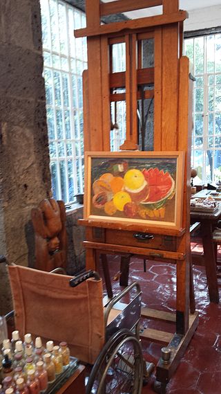 Kahlo’s wheelchair and adjustable easel in La Casa Azul, with one of her still lifes from her final years Author Martinica.ferrara CC BY-SA 4.0