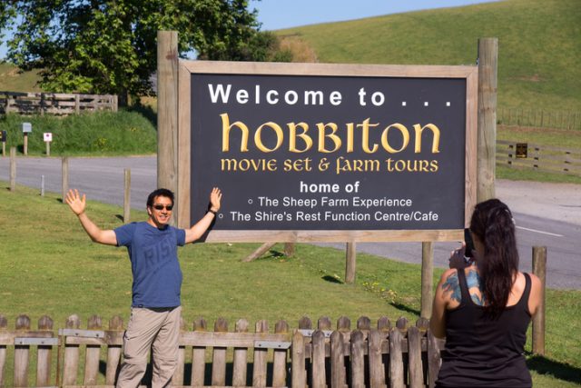 “Matamata, New Zealand – January 7, 2013: A visitor poses for a photo before a welcoming sign to Hobbiton. Hobbiton features an actual movie set of The Shire, used in the filming of the Lord of the Rings trilogy and The Hobbit.”