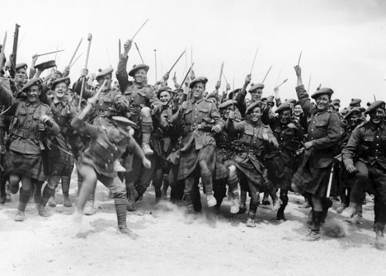 A large group of soldiers, likely South African infantry, having a good time. They are stamping their feet and brandishing anything that comes to hand, from walking sticks to swords. It is all being done in a light-hearted fashion, with most of the men pulling funny faces and smiling. Many of the soldiers are wearing kilts and balmorals. (National Library of Scotland)