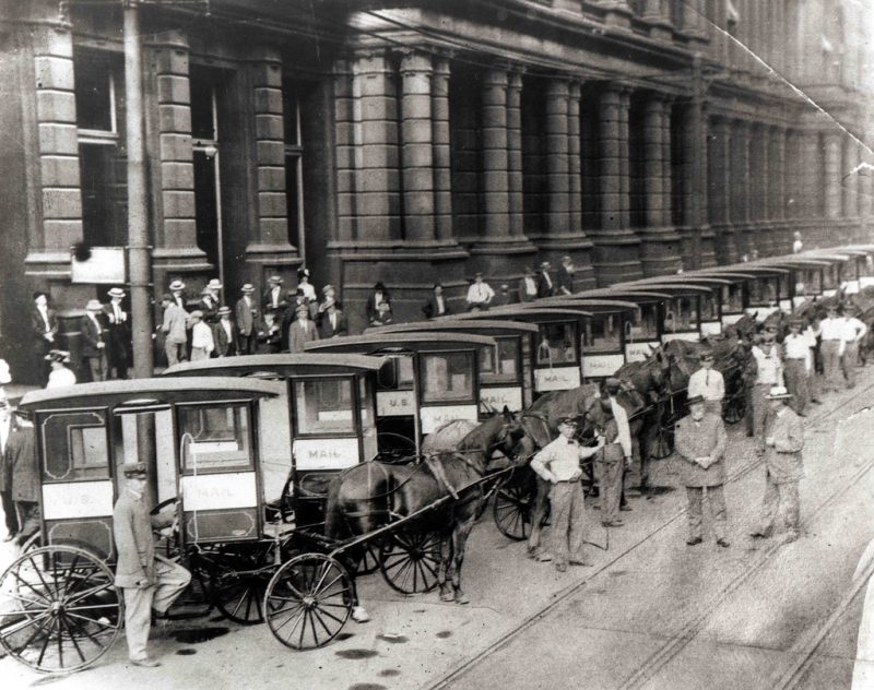 . In an unidentified city, postal officials pose with letter carriers and row of one-man collection mail wagons 1905