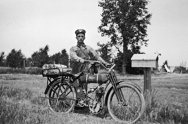 A mail carrier stands with his Wagner 4-11 motorcycle next to a postal box along his route near Newell, South Dakota, 1915