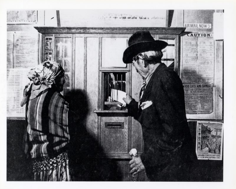 A woman and man pose at the window of a small, fourth-class post office in an unidentified rural town. 1938