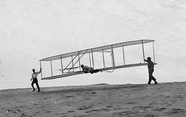 Start of a glide; Wilbur in motion at left holding one end of the glider (rebuilt with single vertical rudder), Orville lying prone in the machine, and Dan Tate at right, in Kitty Hawk, North Carolina, on October 10, 1902.