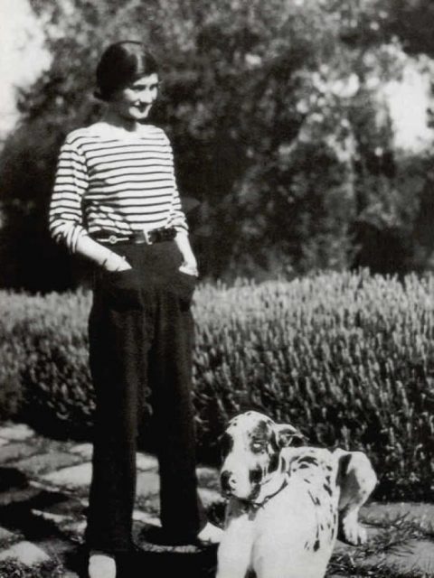 black and white photo of Coco Chanel wearing pants with pockets and a striped shirt