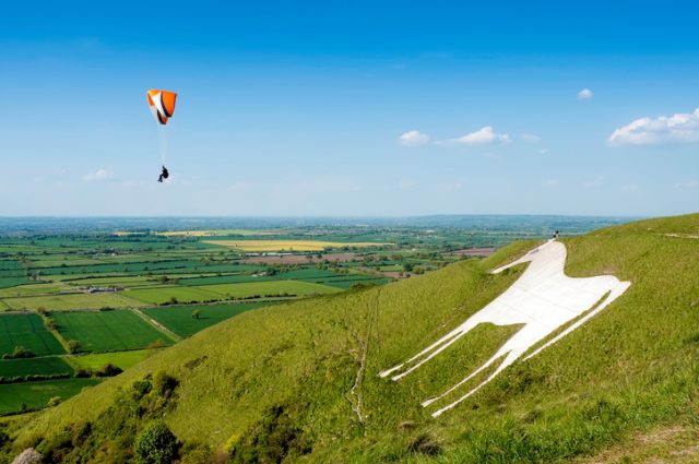 A wide angle view of a paraglider airborne over the famous landmark of the Westbury White Horse hill figure in Wiltshire. Cut into the hillside in 1778, this hill carving replaced an older horse that was possibly cut to commemorate King Alfred’s nearby victory over the Vikings.