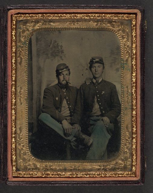 Two unidentified soldiers in Union uniforms in front of painted backdrop showing trees.