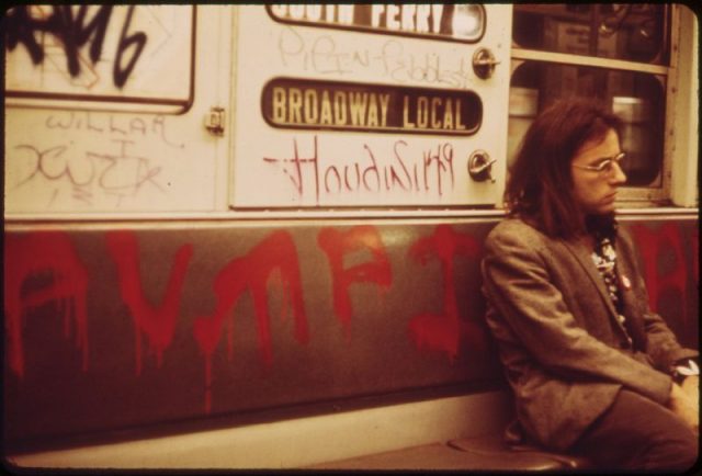 A man sits on a seat on a subway car which has been extensively marked with graffiti, 1973.