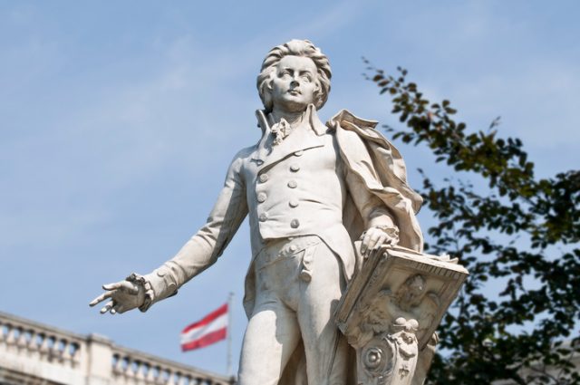 Statue of Wolfgang Amadeus Mozart, located in the Burggarten in Vienna. In the background the Austrian flag on the Austrian National Library.
