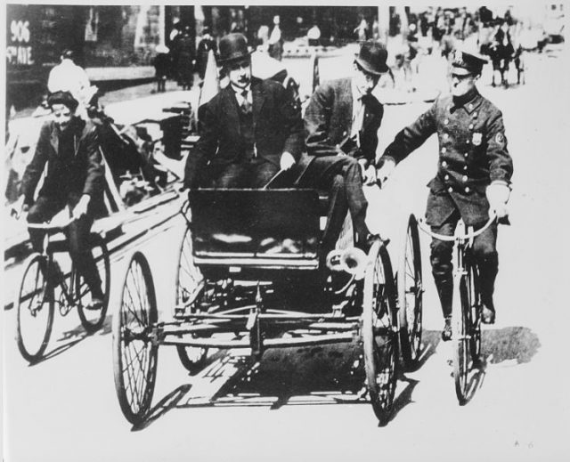 Traffic violator driving a 1900-vintage car being stopped by a policeman on a bicycle.