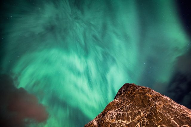 Aurora borealis (Northern lights) dancing in the night sky above a steep mountain peak.