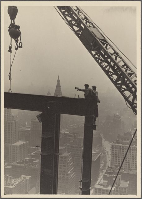 Two construction workers at the corner of two steel beams, pointing to the left.