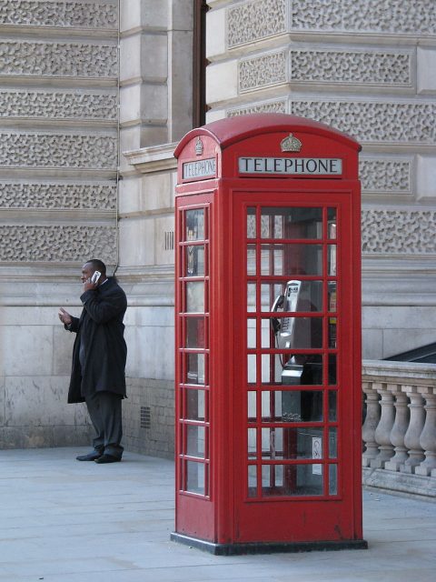 A man talks on his mobile phone while standing near a conventional telephone box, which stands empty. Photo by McKay Savage CC BY 2.0