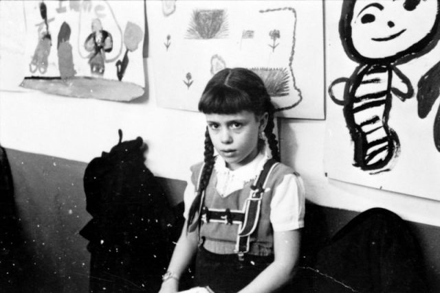 Young girl, half-length portrait, standing against wall displaying art work, in classroom in Chicago, Illinois. Photography by S. Kubrick.
