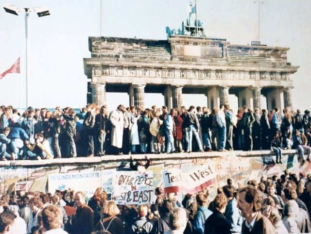The fall of the Berlin Wall, 1989. East and West Germans stand on top of the wall at the Brandenburg Gate in the days before it was torn down. Photo by Lear 21 CC BY SA 3.0