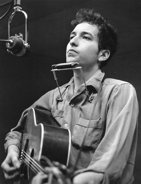 Bob Dylan playing the guitar in front of a microphone, with a harmonica around his neck