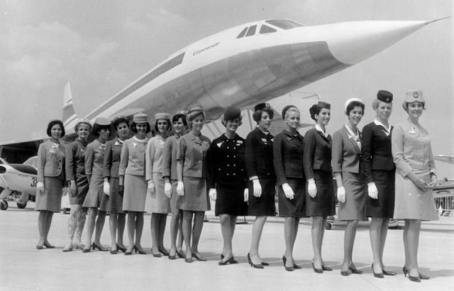 A line-up of some of the air stewardesses who attend to passengers on board the supersonic jet the 'Concorde', each one from a different airline. They are standing in front of a scale model of the aircraft. 
