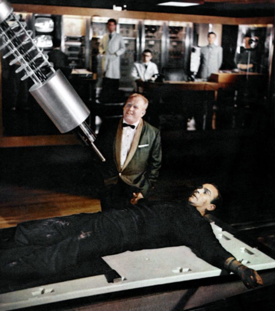 Auric Goldfinger watching while James Bond is strapped to a metal table with a laser above him