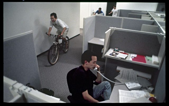 A man sitting in a cubicle while another cycles past him