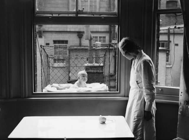 A nanny supervising a baby suspended in a wire cage attached to the outside of a high tenement block window. (Photo Credit: Reg Speller/Fox Photos/Getty Images)