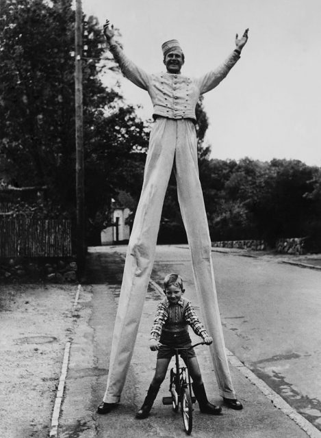 Child sitting on a bicycle below a man on stilts